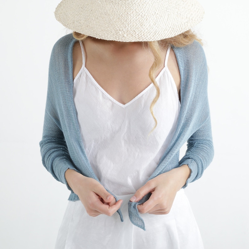 Op de afbeelding: Een lichtblauwe gebreide omslagdoek met lange mouwen. De omslagdoek is aan de voorkant vastgebonden en wordt over een witte camisole gedragen. Een strohoed wordt op het hoofd gedragen.