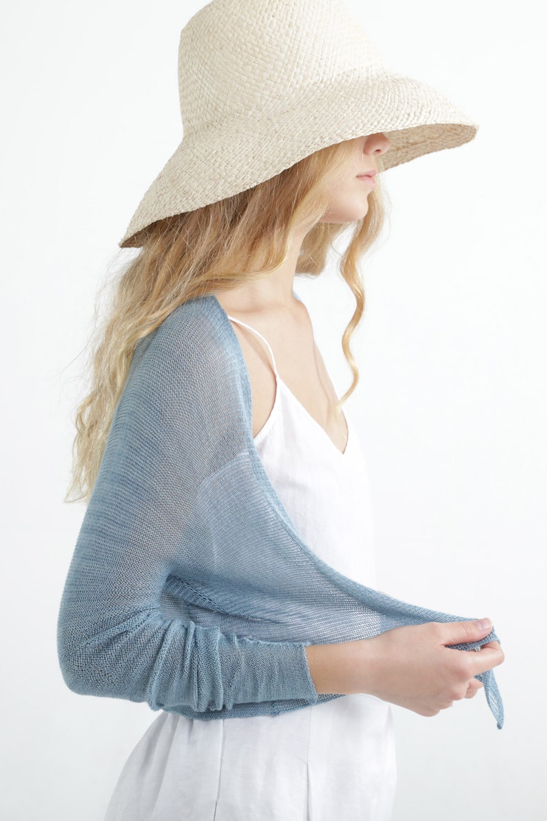 Op de afbeelding: Een vrouw draagt een lichtblauwe, doorschijnende, lange mouwen shrug over een witte top. Ze draagt ook een breedgerande, strokleurige hoed. De outfit is eenvoudig en elegant, met de nadruk op natuurlijke materialen en lichte kleuren.