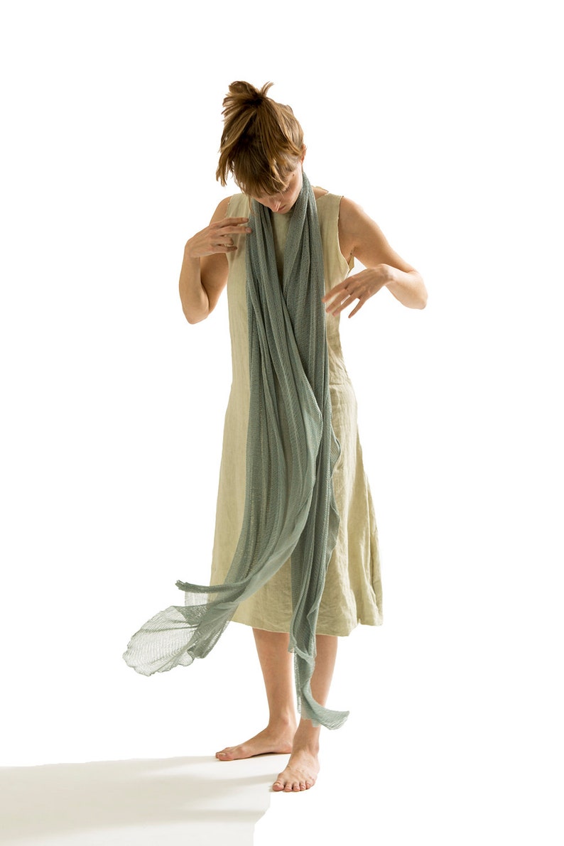 Puede incluir: Una mujer lleva un vestido de lino beige claro con una larga y fluida bufanda verde claro drapeada alrededor de su cuello. La bufanda se mueve con el viento.