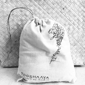 May include: A natural-colored drawstring bag with a black floral design and the text "TOOSH&Lambda;&Lambda;YA Touched by Nature". The bag is made of a soft, textured fabric and is tied at the top with a thin, dark string.