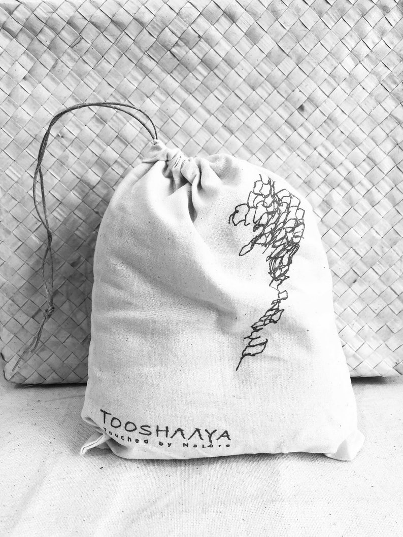 May include: A natural-colored drawstring bag with a black floral design and the text "TOOSH&Lambda;&Lambda;YA Touched by Nature". The bag is made of a soft, textured fabric and is tied at the top with a thin, dark string.