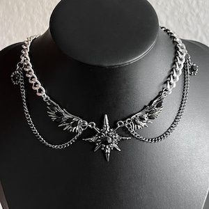 Könnte beinhalten: Eine silberfarbene Metallhalskette mit einem zentralen sternförmigen Anhänger mit schwarzem Stein, flankiert von zwei flügelförmigen Anhängern, die mit schwarzen Kristallen verziert sind. Das Design umfasst eine dicke Panzerkette und dünne Ketten.