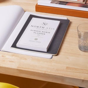 May include: A white North Axis School Nurse Planner book with black text and a logo, resting on a black notebook and an open white notebook. A clear glass of water with seeds is positioned to the right of the books on a light wood surface.