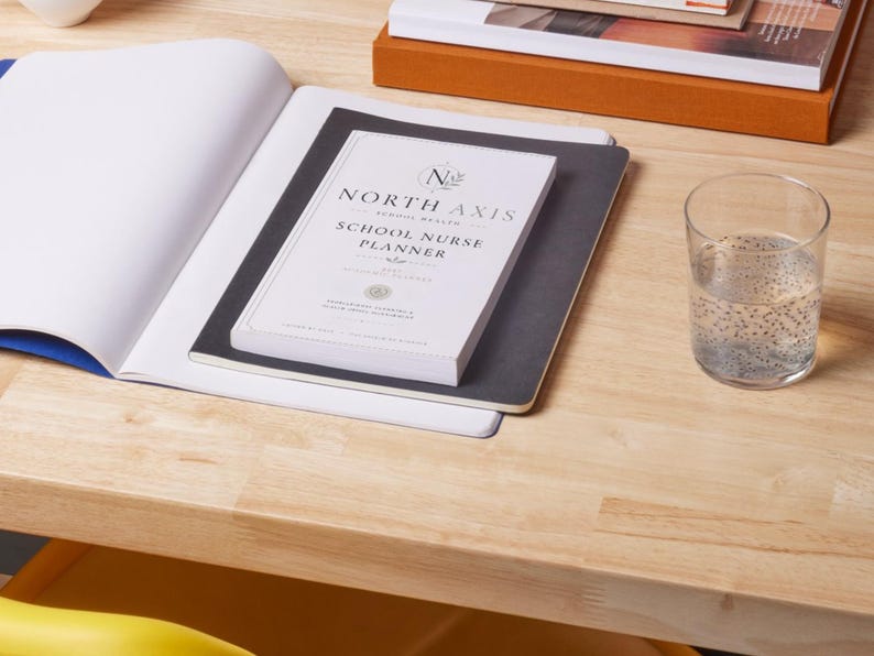 May include: A white North Axis School Nurse Planner book with black text and a logo, resting on a black notebook and an open white notebook. A clear glass of water with seeds is positioned to the right of the books on a light wood surface.