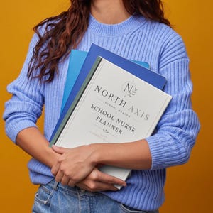 May include: A light blue ribbed sweater and blue jeans holding a stack of notebooks. The top notebook is white with the text North Axis School Nurse Planner, featuring a circular logo with the letter N and a laurel wreath design element.
