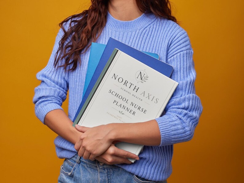 May include: A light blue ribbed sweater and blue jeans holding a stack of notebooks. The top notebook is white with the text North Axis School Nurse Planner, featuring a circular logo with the letter N and a laurel wreath design element.