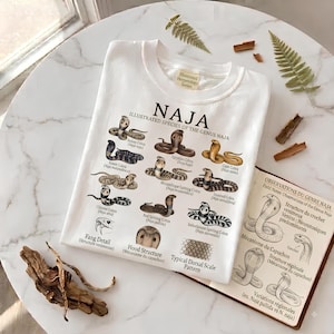 Op de afbeelding: Een wit t-shirt met een gedetailleerde wetenschappelijke illustratie van verschillende Naja-cobra-soorten. De afbeelding bevat gelabelde tekeningen van slangen, slagtanddetails, kapstructuren en schubpatronen met tekst.