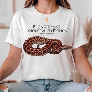 Puede incluir: Una camiseta blanca de manga corta con un gráfico de una pitón de cola corta de Brongersma enroscada con patrones marrones y bronceados. El texto "BRONGERSMA'S SHORT-TAILED PYTHON" y "Python brongersmai" está impreso en negro sobre la serpiente.