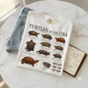 Peut inclure: Un t-shirt blanc avec le texte "TURTLES OF THE USA" et "Illustrated common and diverse species", présentant des illustrations colorées détaillées de diverses espèces de tortues, notamment la tortue peinte, la tortue à tempes rouges et la tortue serpentine.