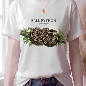 Pu&ograve; includere: Una t-shirt bianca a maniche corte con la grafica di un pitone reale avvolto su un ramo con foglie verdi. Il testo BALL PYTHON (Python regius) &egrave; stampato sopra il serpente. &Egrave; indossata una sottile collana a catena con un ciondolo.