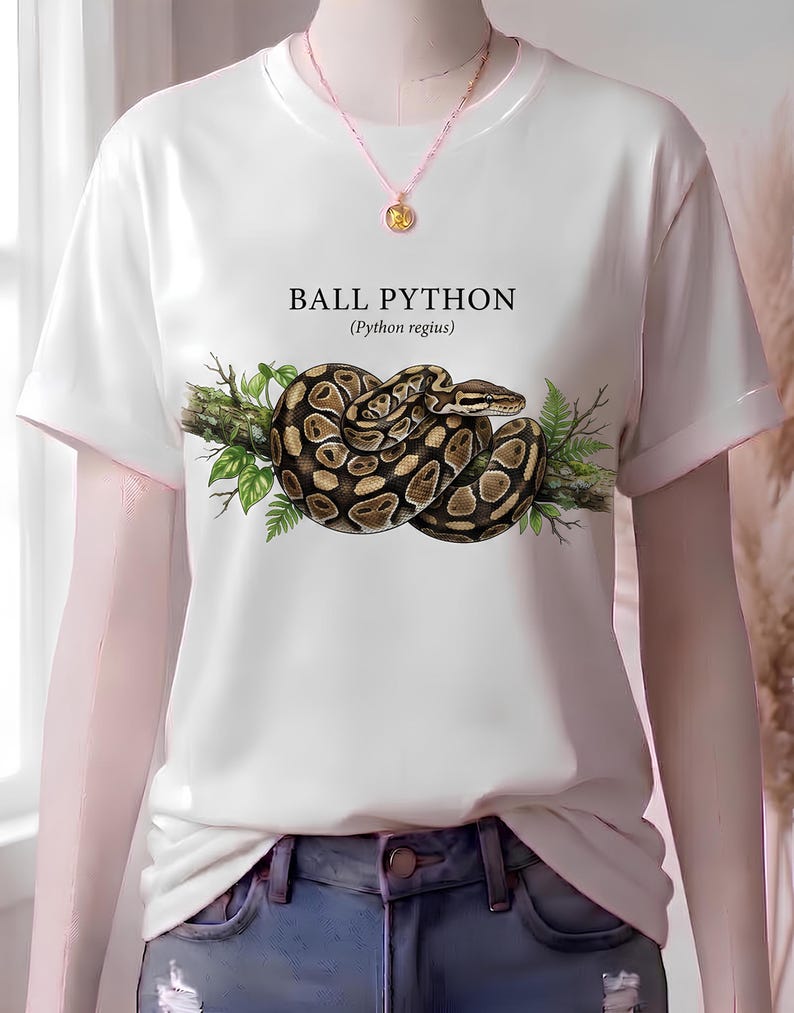Pu&ograve; includere: Una t-shirt bianca a maniche corte con la grafica di un pitone reale avvolto su un ramo con foglie verdi. Il testo BALL PYTHON (Python regius) &egrave; stampato sopra il serpente. &Egrave; indossata una sottile collana a catena con un ciondolo.