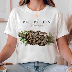 Puede incluir: Una camiseta blanca de manga corta con un gráfico de una pitón bola enroscada en una rama de árbol con hojas verdes. El texto "BALL PYTHON" y "(Python regius)" está impreso en negro sobre la ilustración de la serpiente al frente.