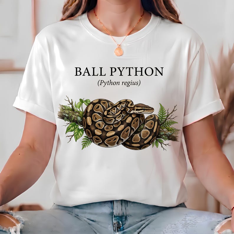 Pu&ograve; includere: Una t-shirt bianca a maniche corte con la grafica di un pitone reale avvolto su un ramo d'albero con foglie verdi. Il testo "BALL PYTHON" e "(Python regius)" &egrave; stampato in nero sopra l'illustrazione del serpente sul davanti.