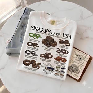 Puede incluir: Una camiseta blanca con un gráfico titulado SNAKES OF THE USA que muestra ilustraciones de varias especies de serpientes, incluyendo la serpiente de cascabel diamantada, la culebra rayada y la serpiente real de California, junto a una pequeña tarjeta informativa.