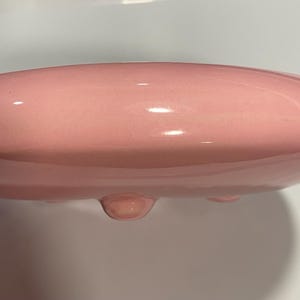 May include: A smooth, glossy, light pink ceramic bowl with a rounded, disc-like shape. The object features a curved, solid surface and small, rounded feet visible along the bottom edge, providing a stable base for the circular, bowl-like structure.