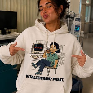 May include: A cream-colored hooded sweatshirt featuring a graphic illustration of a medical professional sitting in a chair, holding a coffee mug, with a monitor displaying NO SIGNAL and the text VITALZEICHEN? PASST. printed below the graphic.