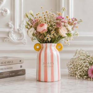 May include: A ceramic vase with vertical pink and white stripes and two yellow circular handles. The front features the engraved text Jack & Lily 03.25.2026. The vase is filled with a colorful arrangement of daisies, pink roses, and baby's breath.