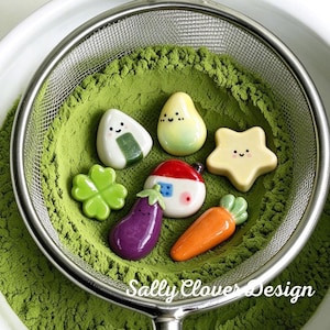 Op de afbeelding: Een verzameling kleine keramische bedels in de vorm van een onigiri, peer, ster, klavertjevier, huisje, aubergine en wortel, rustend op een bedje van groene matchapoeder in een metalen zeef met de tekst Sally Clover Design.