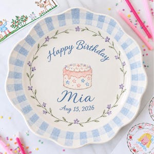 May include: A round ceramic plate with a scalloped edge and a light blue and white gingham border. The centre features a hand-painted illustration of a cake with blue and pink stars, surrounded by a floral vine, text reading Happy Birthday Mia Aug 15, 2026.