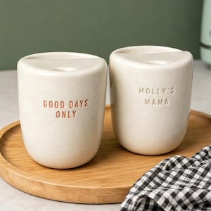 May include: Two off-white ceramic cups with a smooth finish and a semi-circular top opening. One cup features the text "GOOD DAYS ONLY" in orange, while the other displays the text "MOLLY'S MAMA" in a debossed style on the front surface.