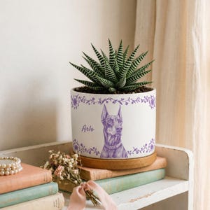 Può includere: Un vaso da fiori in ceramica bianca con un'illustrazione viola di un cane Dobermann e il nome Arlo stampato in corsivo viola. Il vaso è decorato con un bordo di viticci floreali viola e contiene una pianta succulenta a strisce verdi e bianche.