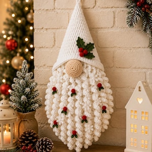 May include: A white crocheted gnome wall hanging featuring a tall pointed hat, a round beige nose, and a long textured beard adorned with small red berries and green leaves. The gnome has a loop at the top for hanging and a white crochet finish.