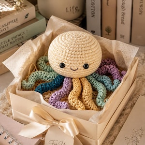 Peut inclure: Une pieuvre en crochet de couleur crème, ronde, avec des yeux de sécurité noirs et un sourire brodé. La pieuvre possède plusieurs tentacules dans des tons vert menthe, bleu sarcelle, lavande et jaune, placée dans une boîte cadeau carrée.