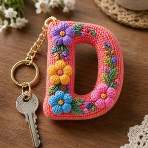 Puede incluir: Un llavero con la letra D tejida a ganchillo en color coral, decorado con bordados florales en morado, amarillo, azul y rosa con hojas verdes. La letra está unida a una cadena de metal dorado y a una llave de metal de color plateado.
