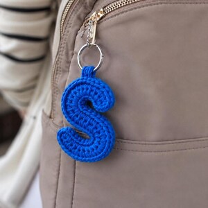 May include: A bright blue crochet letter S keychain attached to a silver metal ring. The letter is crafted with a dense, textured yarn stitch pattern and hangs from the zipper pull of a tan-colored backpack, showcasing a vibrant, solid color.