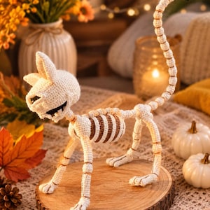 May include: A crocheted cat skeleton figurine crafted from off-white yarn with brown rib cage detailing. The figure features a long, upright tail, pointed ears, and an open mouth design, standing on a circular wooden base with visible grain.