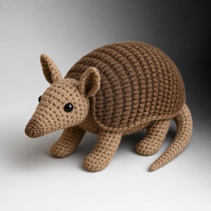Puede incluir: Un juguete de armadillo tejido a ganchillo a mano con cabeza, patas y cola de color tostado claro y un caparazón redondeado y segmentado de color marrón oscuro. La criatura tiene dos orejas pequeñas erguidas y dos ojos negros redondos.