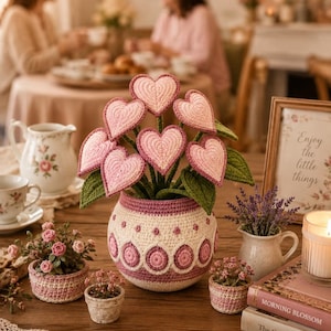 May include: A crochet flower arrangement featuring seven pink heart-shaped blossoms with green leaves, housed in a round, cream and pink patterned crochet pot, accompanied by two smaller crochet pots containing miniature pink flowers.