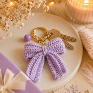 May include: A lavender crochet bow keychain featuring a gold-toned metal ring, a small purple bead accent, and two silver-toned keys attached to the keyring. The bow is constructed from yarn with a visible ribbed texture and a central knotted detail.