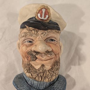 May include: A ceramic bust of a bearded sailor wearing a cream-colored captain's hat with a black brim and a red anchor emblem. The figure has a textured blue sweater, a weathered face with deep wrinkles, and a brown cigar held in the mouth.