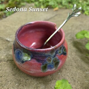 May include: A small, round, red ceramic bowl with a silver spoon. The bowl has a textured surface with blue and green accents. The text "Sedona Sunset" is at the top.