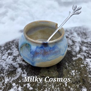 May include: A small, round ceramic bowl with a blue and tan glaze, and a silver spoon with a leaf design. The bowl is labeled "Milky Cosmos" in white text.
