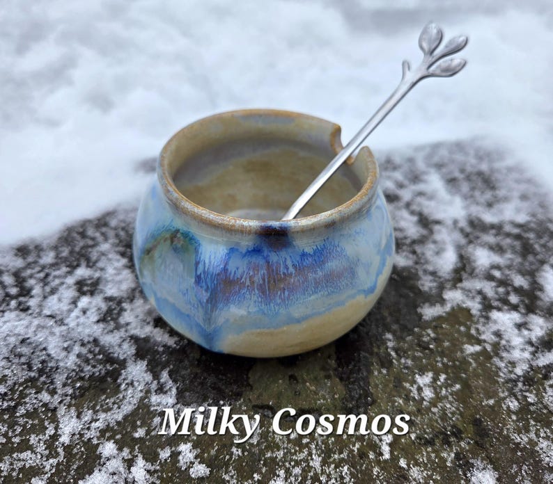 May include: A small, round ceramic bowl with a blue and tan glaze, and a silver spoon with a leaf design. The bowl is labeled "Milky Cosmos" in white text.