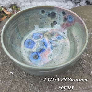 May include: A handmade ceramic bowl with a textured, glazed surface. The bowl features a blend of blue, green, and pink hues with a speckled pattern. The text "4 1/4x1 23 Summer Forest" is printed on the bottom of the bowl.