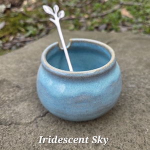 May include: A small, round, light blue ceramic jar with a matching spoon. The jar has a slightly textured surface and a light brown rim. The spoon is white and has a decorative leaf design on the handle. The text "Iridescent Sky" is at the bottom.