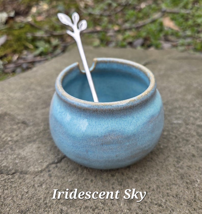May include: A small, round, light blue ceramic jar with a matching spoon. The jar has a slightly textured surface and a light brown rim. The spoon is white and has a decorative leaf design on the handle. The text "Iridescent Sky" is at the bottom.