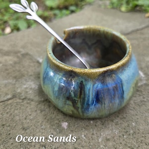 May include: A ceramic bowl with a blue and green glaze, featuring a silver spoon with a leaf design. The bowl has a rounded shape and a golden rim. The text "Ocean Sands" is at the bottom of the image.