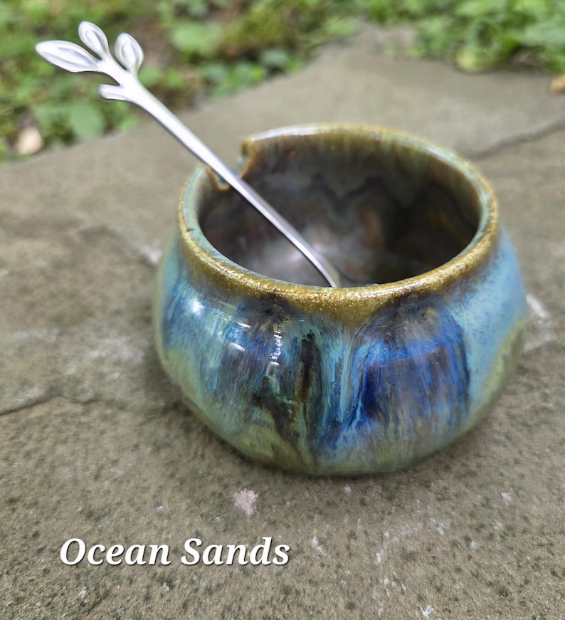 May include: A ceramic bowl with a blue and green glaze, featuring a silver spoon with a leaf design. The bowl has a rounded shape and a golden rim. The text "Ocean Sands" is at the bottom of the image.