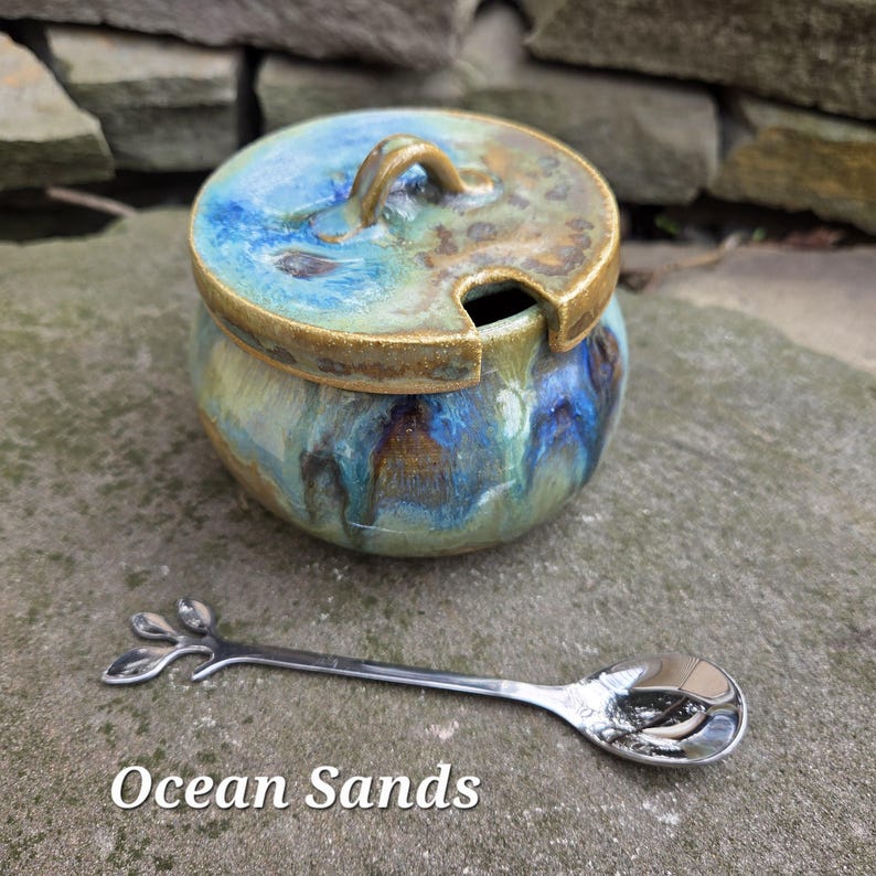May include: A ceramic jar with a lid, featuring blue, green, and brown glaze. A small, silver spoon with a leaf design rests beside the jar. The text "Ocean Sands" is at the bottom of the image.