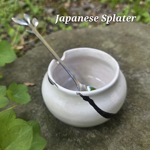 May include: A small, white ceramic bowl with a silver spoon. The bowl has a black accent line and the spoon has a leaf design. The text "Japanese Splater" is above the bowl.