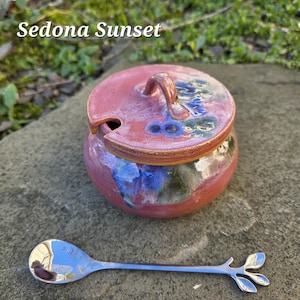 May include: A ceramic jar with a lid, in shades of red, blue, and green, with the text "Sedona Sunset". A silver spoon with a leaf design is next to the jar. The jar has a small handle on the lid.