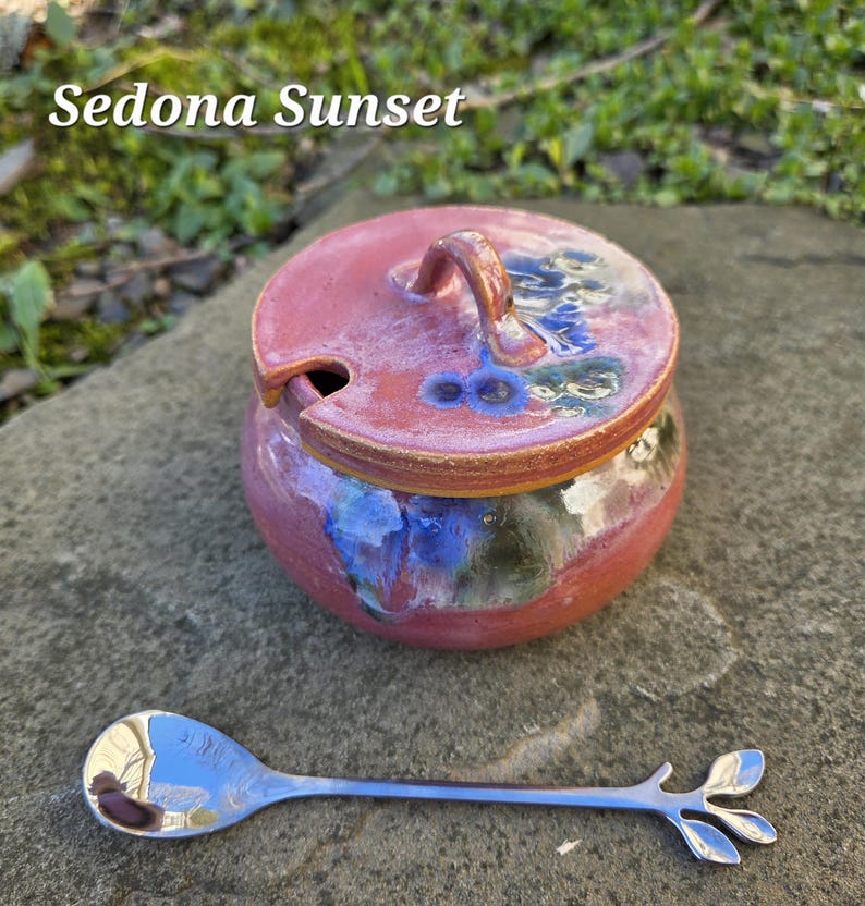 May include: A ceramic jar with a lid, in shades of red, blue, and green, with the text "Sedona Sunset". A silver spoon with a leaf design is next to the jar. The jar has a small handle on the lid.