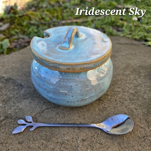 May include: A ceramic jar with a lid, glazed in shades of blue and brown, with the text "Iridescent Sky". A silver spoon with a leaf design rests beside the jar. The jar is round with a handle on the lid.