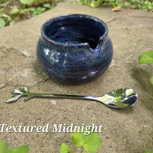 May include: A textured, midnight blue ceramic bowl with a spout, paired with a silver spoon featuring a leaf design. The spoon rests on a stone surface. The text "Textured Midnight" is at the bottom.