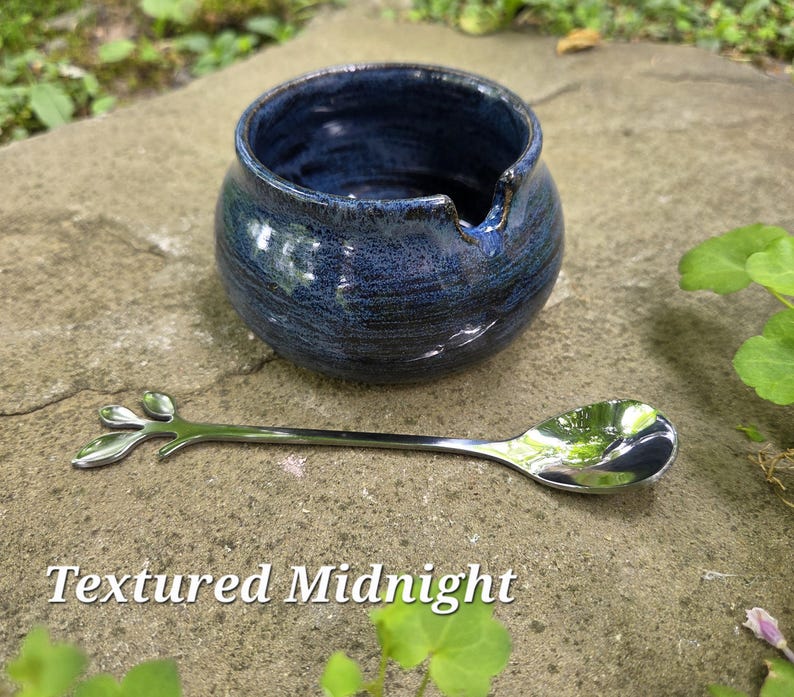 May include: A textured, midnight blue ceramic bowl with a spout, paired with a silver spoon featuring a leaf design. The spoon rests on a stone surface. The text "Textured Midnight" is at the bottom.