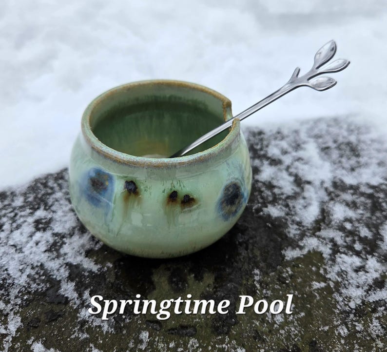 May include: A small, round ceramic bowl with a light green glaze and blue accents. A silver spoon with a leaf-like design rests inside the bowl. The text "Springtime Pool" is at the bottom of the image.